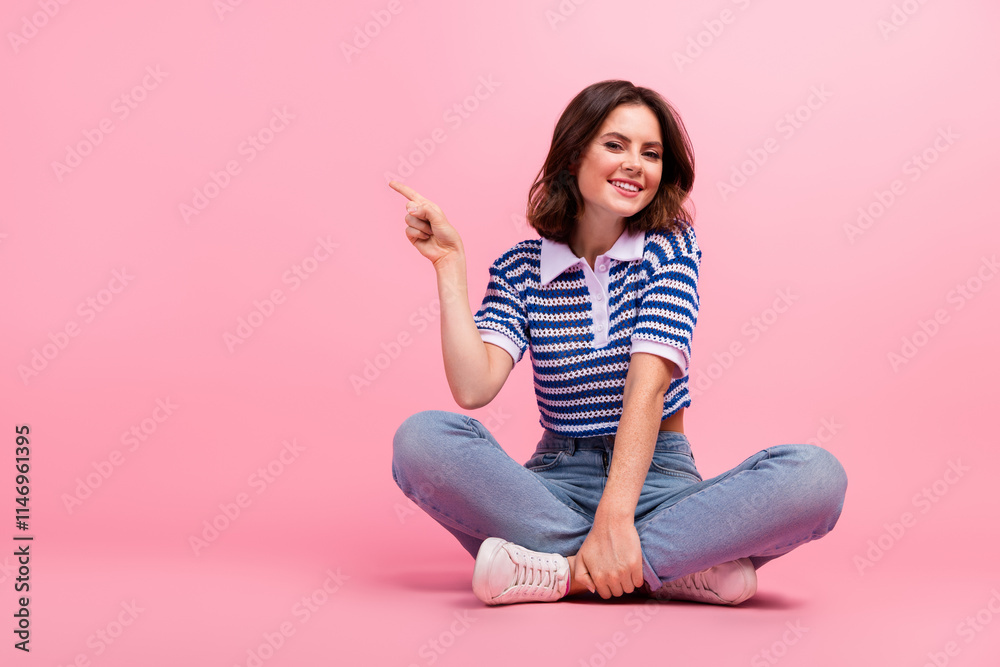 Charming young girl in casual wear pointing and smiling while sitting on a pink background Charming young girl in casual wear pointing and smiling while sitting on a pink background