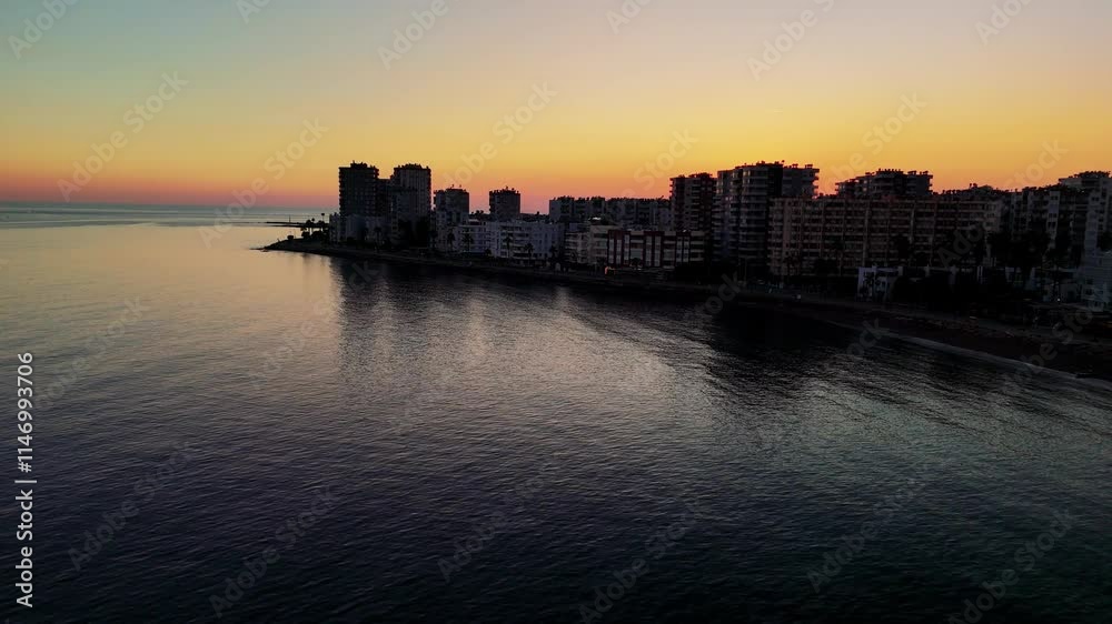Warm golden light enveloping mersin's coastline, casting elongated shadows from urban structures onto serene mediterranean waters during twilight hours Warm golden light enveloping mersin's coastline, casting elongated shadows from urban structures onto serene mediterranean waters during twilight hours