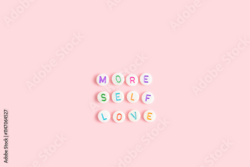 More self love. Quote made of white round beads with multicolored letters on a pink background.