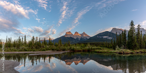 Three Sisters in Canmore seen at golden hour, sunset on blue sky day, afternoon with calm, peaceful reflection in water below famous, tourist, tourism mountains, area summer.	