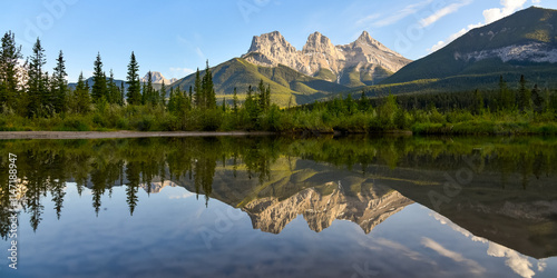 Three Sisters in Canmore seen at golden hour, sunset on blue sky day, afternoon with calm, peaceful reflection in water below famous, tourist, tourism mountains, area summer.	