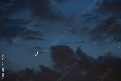 Fototapeta Naklejka Na Ścianę i Meble -  A crescent moon dances with Venus on a blue backdrop as night fall slowly blankets an evening sky dotted with wispy clouds showing hues of purple and orange.