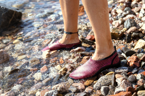 Feet of a young woman wearing water shoes standing on a stony sea beach close up. Protection from sharp stones