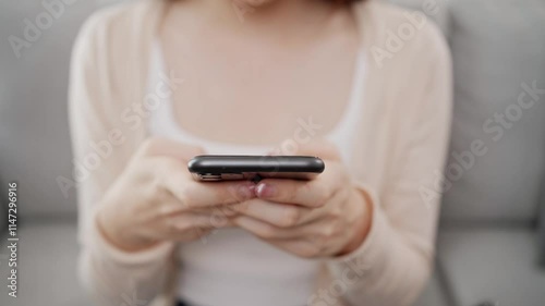 Close up video of a beautiful and lovely Asian woman using a smartphone, sitting on a sofa in a living room at home. Using technology at home concept.