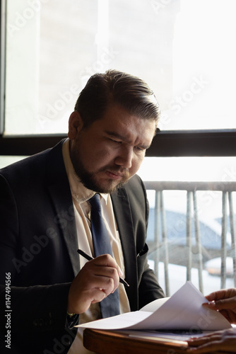 Focused businessman in suit reviewing, signing documents with a pen while sitting in his office with paperwork on his desk, reading a contract or agreement