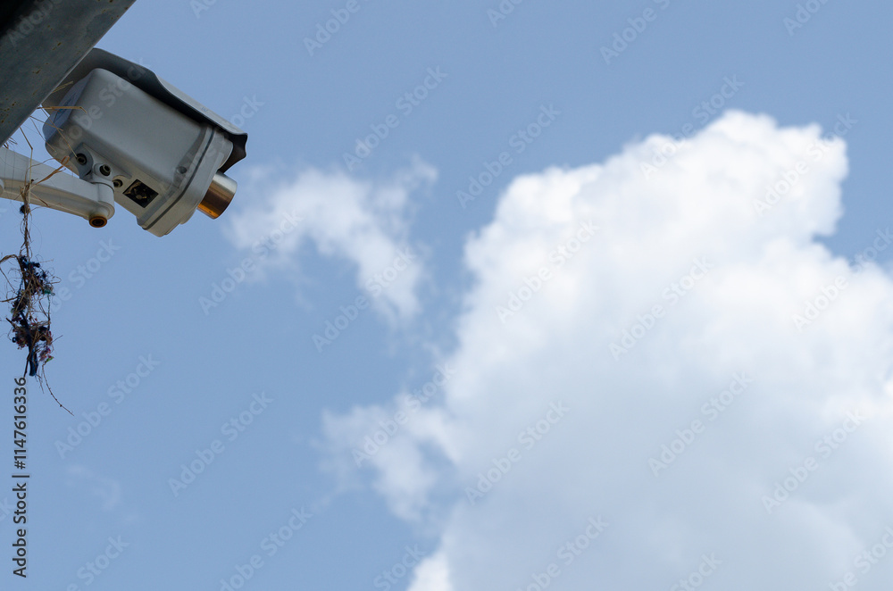 Low-angle, close-up view of a CCTV on a pole against a fluffy cloud and blue sky. Low-angle, close-up view of a CCTV on a pole against a fluffy cloud and blue sky.