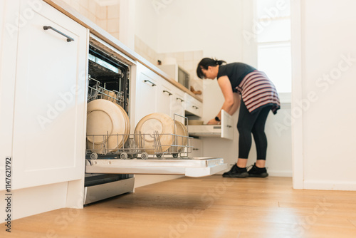 Woman unloading dishwasher in bright kitchen with clean plates and cookware on wooden floor