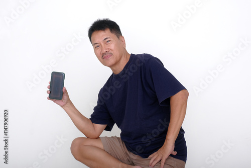 Asian guy in casual clothes, sitting alone on white stool showing messages from his smartphone in his hand photo