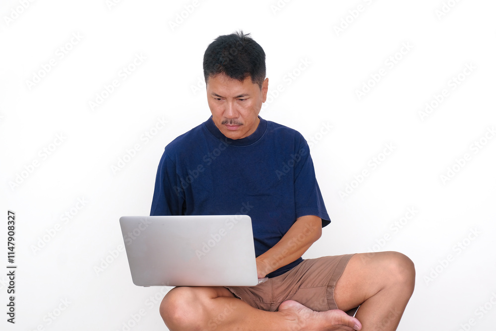 Asian guy in casual clothes, sitting alone on white stool working on his laptop computer Asian guy in casual clothes, sitting alone on white stool working on his laptop computer