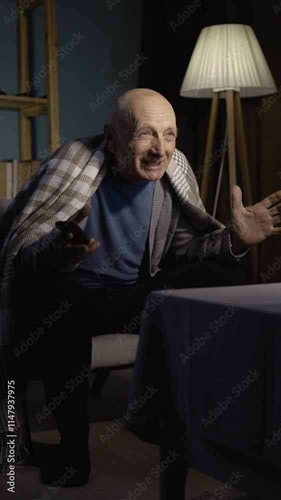 Emotionally senior man comments match watching TV holding remote control at night at home Emotionally senior man comments match watching TV holding remote control at night at home