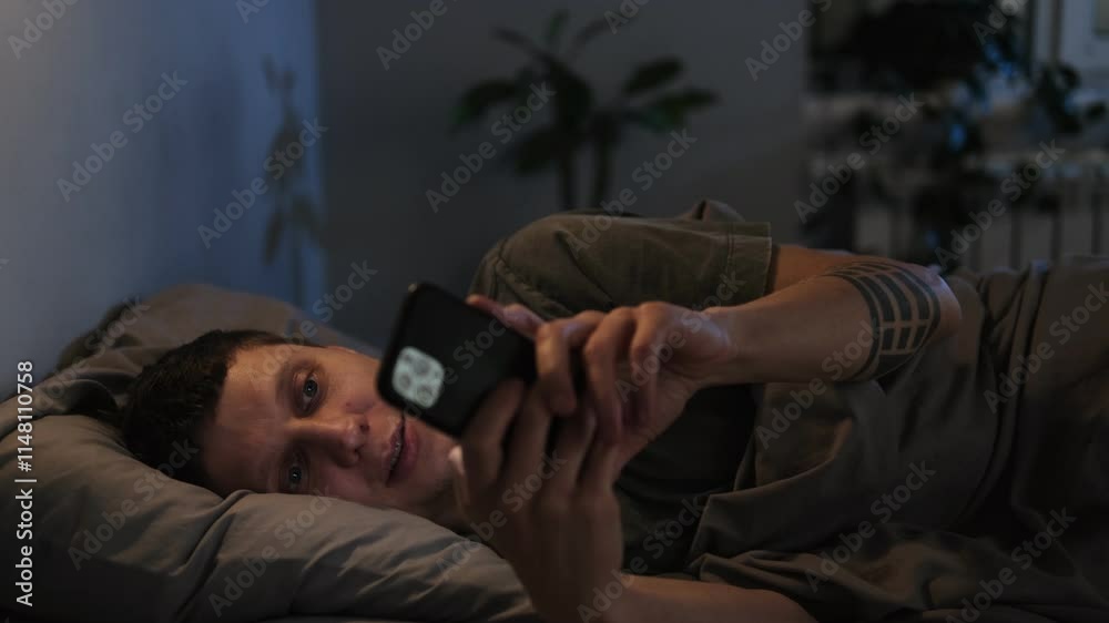 A man lies in a cozy bedroom emotionally scrolling through his phone, unable to tear himself away from the screen. The millennial browses content endlessly in bed until he eventually falls asleep.
