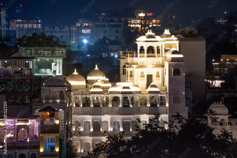 Fototapeta premium Aerial drone shot of brightly lit building exterior with elegant arches, ornate balconies and roof with guests enjoying the evening in Udaipur