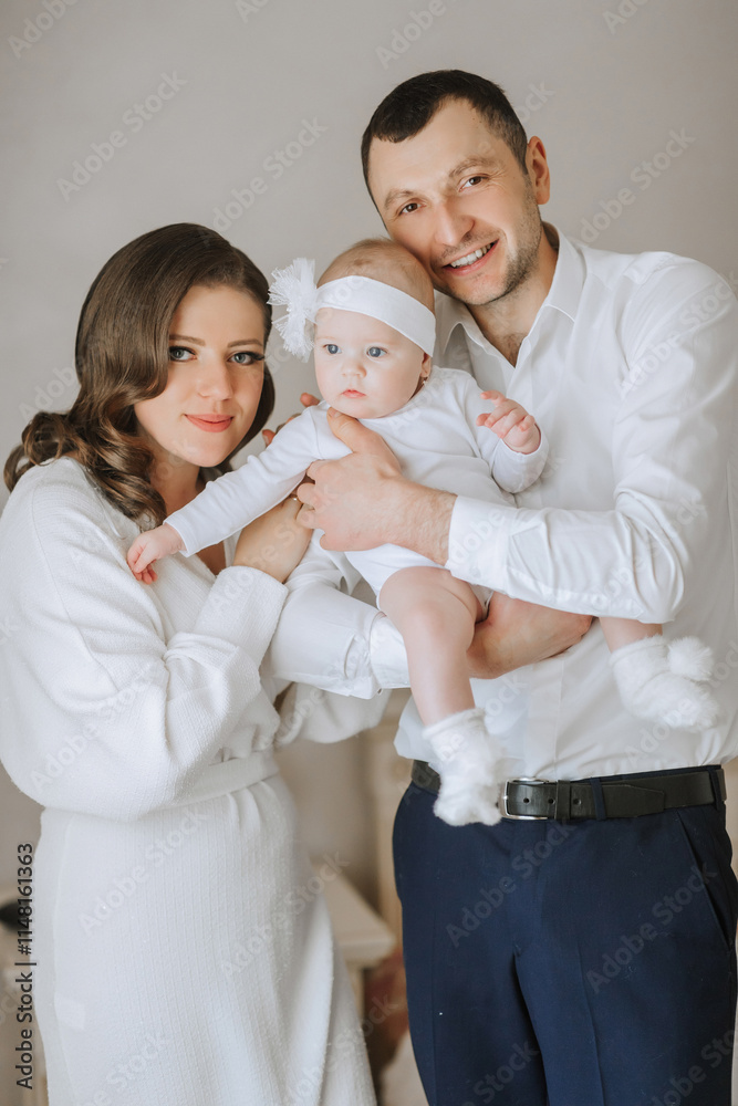 A family of three, a man and two women, are holding a baby. The baby is wearing a white dress and a white headband. The family is smiling and he is happy A family of three, a man and two women, are holding a baby. The baby is wearing a white dress and a white headband. The family is smiling and he is happy