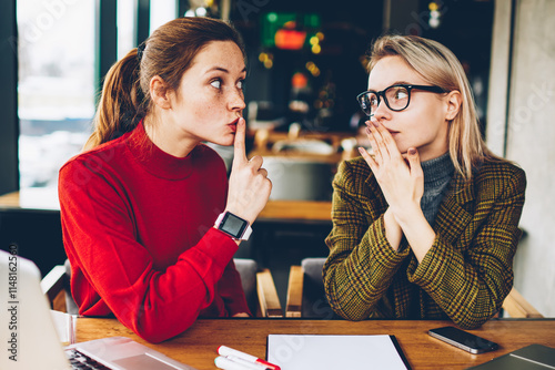 Brunette hipster blogger told secret to wondered colleague and showing sign shh during collaborating at laptop devices in coworking.Excited two best friends gossiping during studying break at netbooks photo