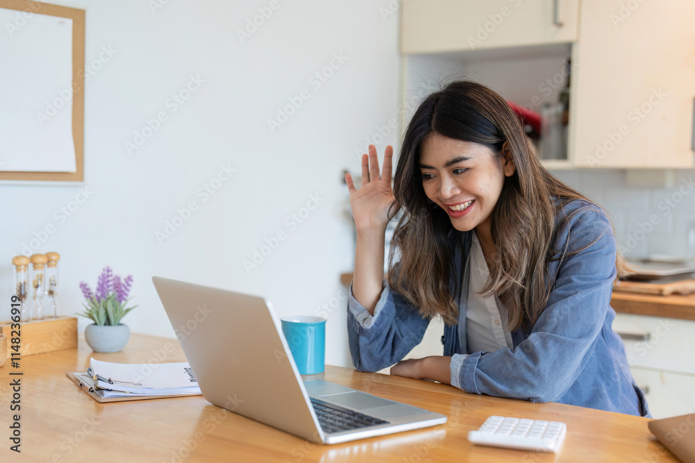 Young Freelance Woman Working from Home in Dining Room, Smiling and Waving at Laptop Screen, Remote Work Concept Young Freelance Woman Working from Home in Dining Room, Smiling and Waving at Laptop Screen, Remote Work Concept