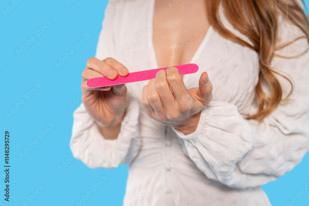 Woman filing her nails with a pink manicure tool against a bright blue background Woman filing her nails with a pink manicure tool against a bright blue background