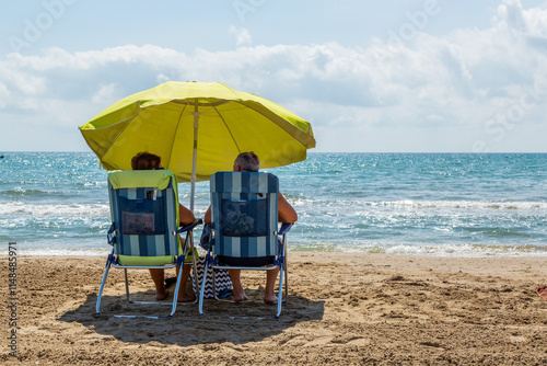 Elderly couple sitting on deck chairs under a parasol by the sea