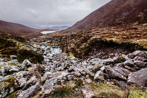 A mountain valley with a small stream running through the center. The valley is filled with rocks and vegetation. There are hills on both sides of the valley. The sky is cloudy.