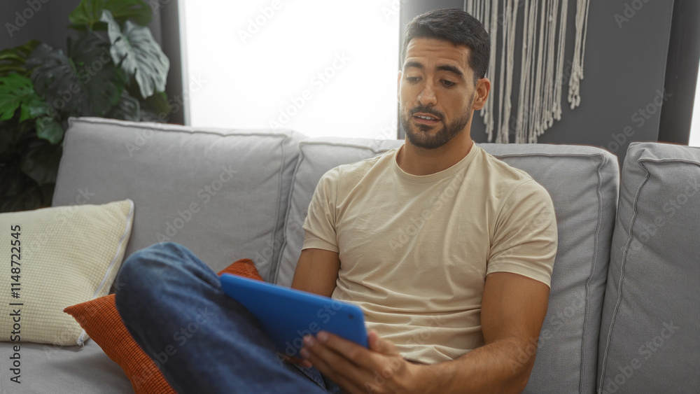 Fototapeta premium Young man indoors at home with beard relaxing on sofa using tablet in cozy living room with pillows and decorative plants enjoying a casual moment in a modern apartment setting.