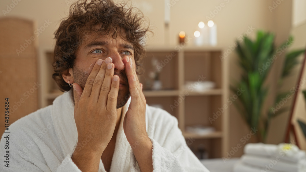 Handsome young man wearing a white robe applying skincare product to his face while sitting in a peaceful spa room Handsome young man wearing a white robe applying skincare product to his face while sitting in a peaceful spa room