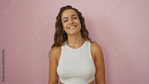 Beautiful young hispanic woman with brunette hair smiling attractively in a white tank top over an isolated pink wall background