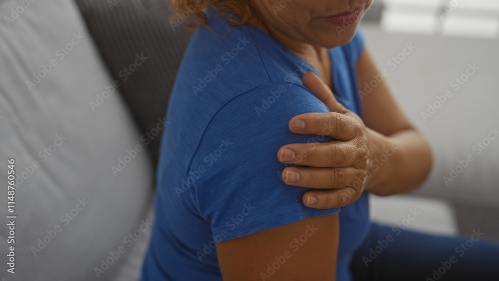A woman holding her shoulder with her hand indoors, seated in a living room, wearing a blue shirt, suggesting pain or discomfort in her arm. A woman holding her shoulder with her hand indoors, seated in a living room, wearing a blue shirt, suggesting pain or discomfort in her arm.