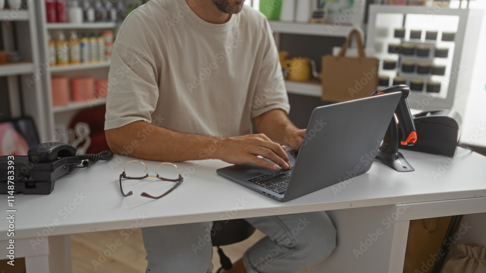 Young man working on laptop in home decor store with shelves of decorations and phone on the table Young man working on laptop in home decor store with shelves of decorations and phone on the table