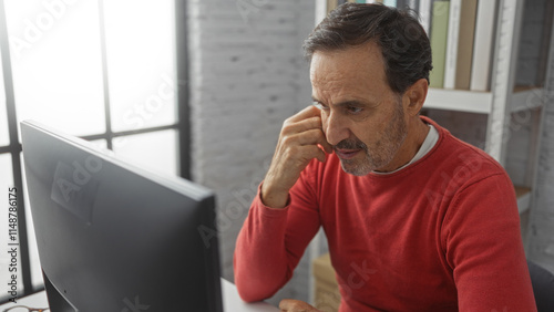 Hispanic man in a red sweater focused on a computer screen in an office setting, emphasizing thoughtful concentration and workplace ambiance.