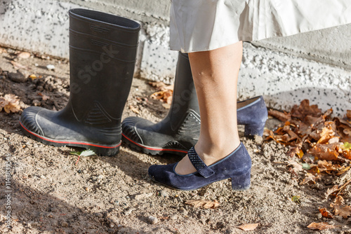 Pair of black Dunlop work boots and a person's blue shoes on dirt and autumn leaves.  Outdoor scene.