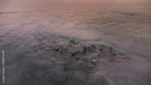 Aerial view captures a small town partially obscured by a thick blanket of mist during a serene sunrise, creating a mystical atmosphere