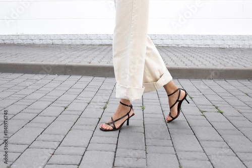 Woman in white denim pants and black heeled sandals walking outside along a city street covered with paving slabs. Trendy elegant casual outfit. Street fashion. Women's legs, without a face.