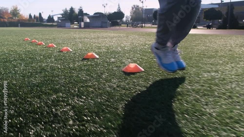 Close up of feet during football training. Athlete in grey pants and blue sneakers, navigating around orange cones on artificial turf
