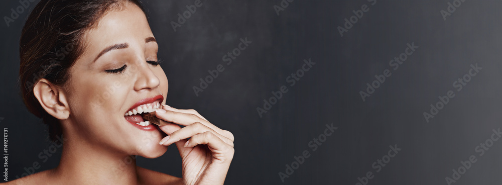 Obraz premium Happy woman, chocolate and bite with candy in studio for unhealthy diet, sweet or dessert on a dark background. Banner, face or female person with cacao, delight or delicious treat on mockup space