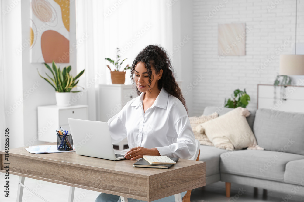 Pretty young African-American woman with laptop sitting at table in home office Pretty young African-American woman with laptop sitting at table in home office