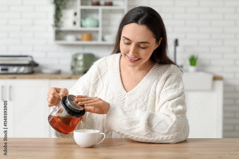 Beautiful young happy woman pouring hot tea from transparent kettle into cup in kitchen Beautiful young happy woman pouring hot tea from transparent kettle into cup in kitchen