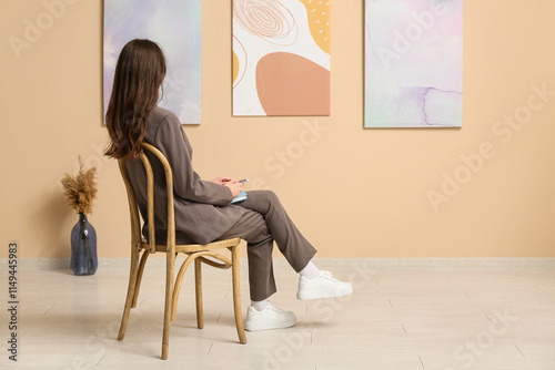 Young woman in chair near paintings on beige wall at art gallery exhibition, ...