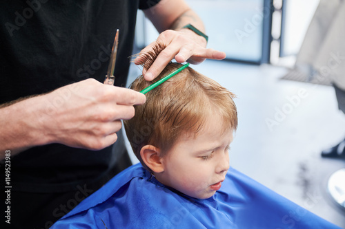 Man barber cutting little boy's hair using comb and scissors. Child getting haircut from adult male, likely barber. Professional hairdresser and cute client at modern barbershop. photo