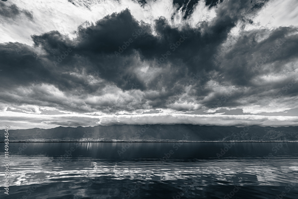 Fototapeta premium Dramatic monochrome landscape of Erhai Lake in Dali, Yunnan, China. Heavy clouds contrast with the serene water, reflecting the mountains and sky. Sunbeams pierce the clouds, creating a breathtaking