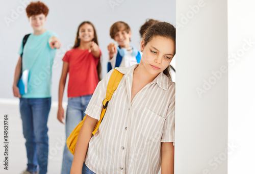 Canvas Print Outcast sad preteen girl suffering from bullying, feeling lonely and excluded, standing alone with group of classmates laughing behind her back and pointing their fingers at her in school corridor