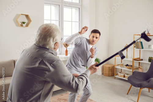 Male patient demonstrates violent behaviour at retirement house. Angry frustrated enraged exasperated hateful aggressive old man raises his crutch to hit young scared terrified fearful nurse assistant