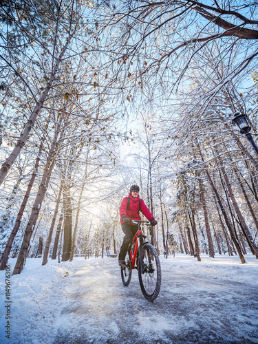 A cyclist is riding through a winter park in the early morning. The man is wearing a red jacket, helmet. Ecological transportation in the city