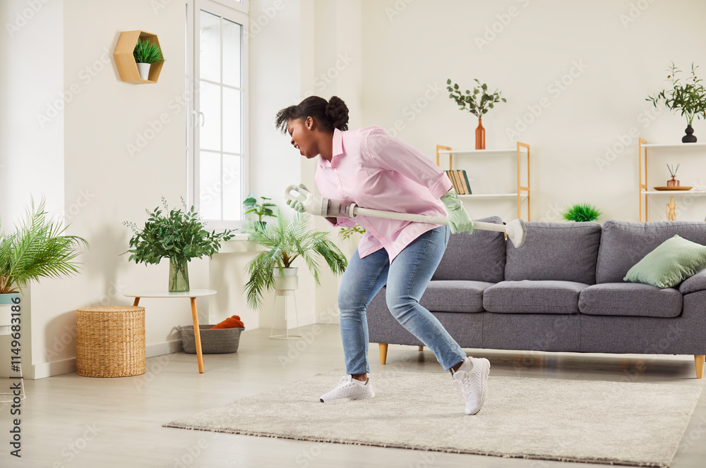 Obraz premium Happy excited funny middle aged african american woman singing at vacuum cleaner hoover cordless tube as microphone, cleaning floor carpet at home, enjoying household activities, vacuuming, having fun
