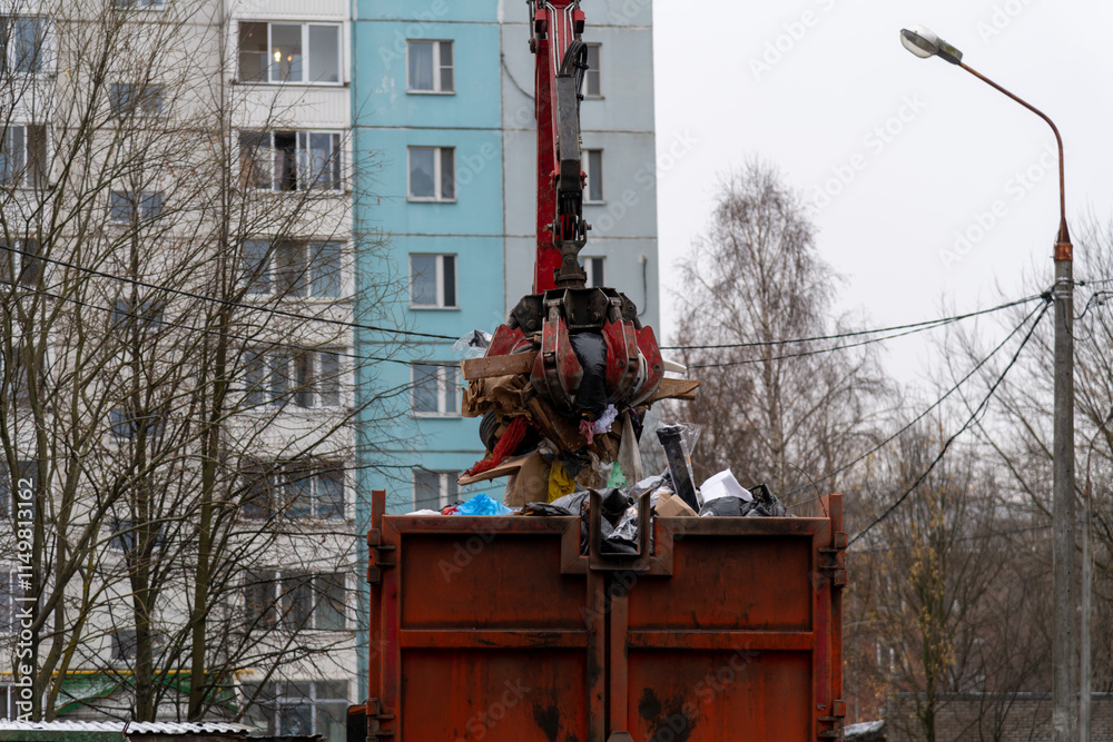 Fototapeta premium Side view of orange machine crane loader manipulator with crab bucket holding bunch of garbage over garbage truck in urban residential district in a cloudy autumn day. Soft focus. Industrial machines.