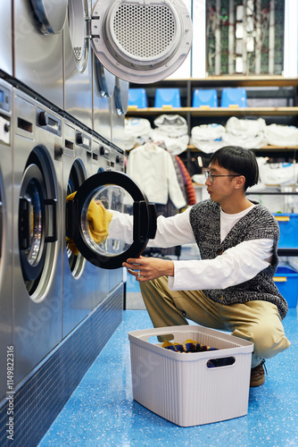 Vertical shot of male Asian customer unloading washing machine putting fresh clean clothes into laundry basket in self service laundry, household chores concept photo