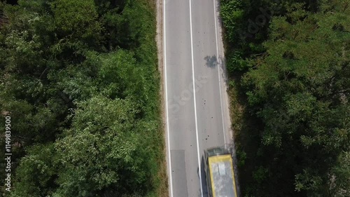 A view of the two-lane road that goes through the forest. A white car passes by, followed by a white-and-yellow bus. Aerial view directly above	