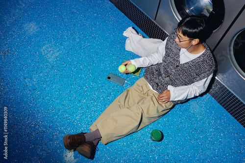 Top view of young man listening to music in headphones while passing time in self service laundry sitting on floor and playing with tennis balls in hand, camera flash, copy space photo