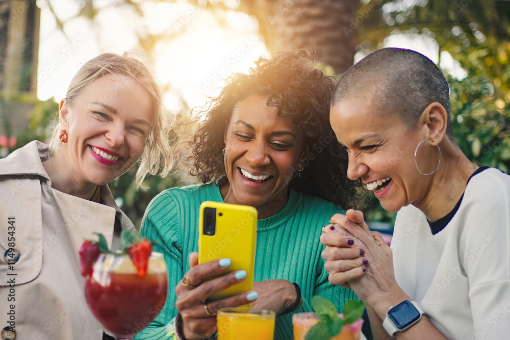 Fototapeta premium Three cheerful multi-ethnic female friends enjoying drinks at a bar, having fun watching something on a mobile phone app