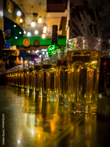 Golden Glow of Celebration. A row of golden-hued glasses aligned on a table in a warmly lit bar, capturing the lively ambiance with blurred lights and shadows in the background.
