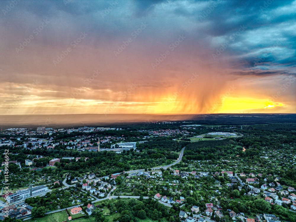 Naklejka premium Sunset Rainstorm Over Polish City. A dramatic aerial view of a sunset rainstorm over a Polish city, with vivid orange and yellow skies contrasting against dark clouds and urban greenery.