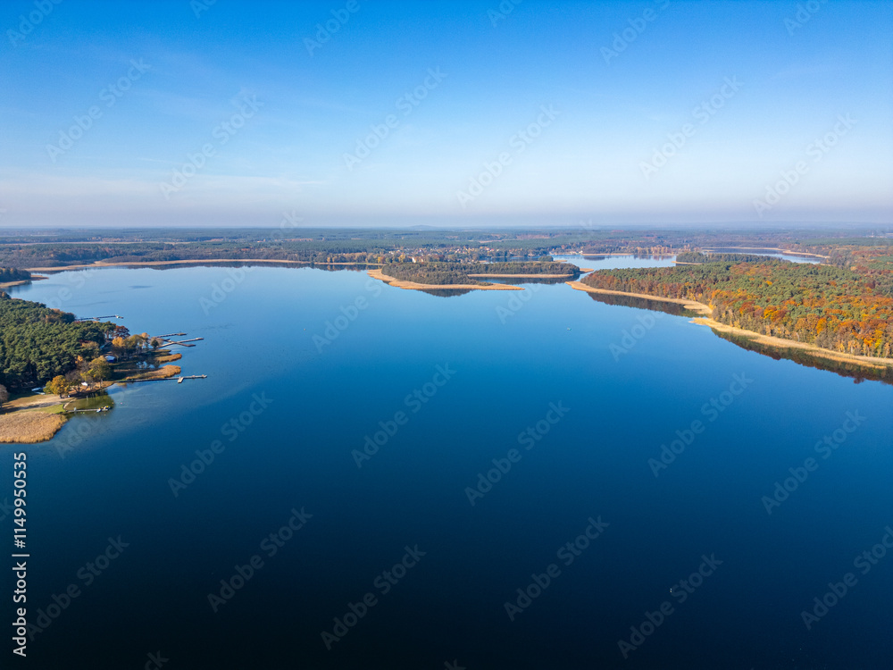 Fototapeta premium A sweeping aerial view of a vast lake surrounded by lush forests with docks and tranquil waters, framed by a clear blue sky.